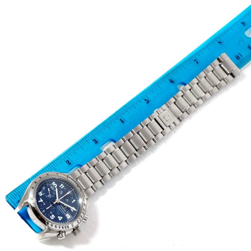 The image shows an Omega Speedmaster Men
s Stainless Steel Blue Dial Men
s Stainless Steel Blue Dial watch facing upward, highlighting its face, chronograph buttons, and stainless steel bracelet next to a blue ruler.