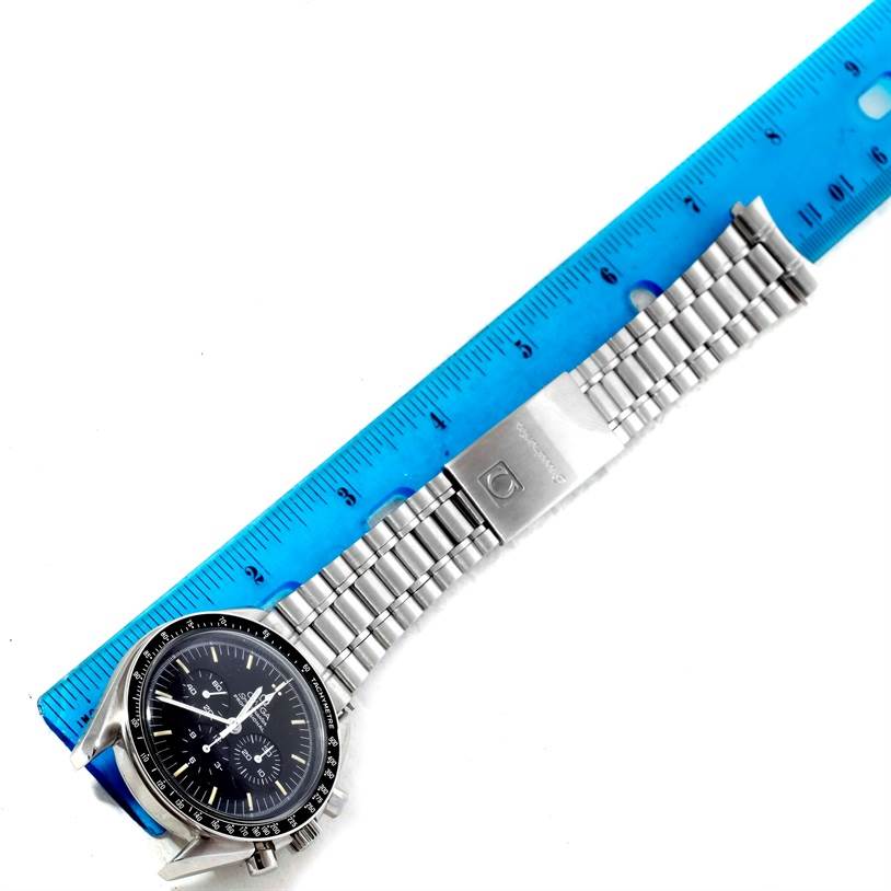 The image shows an Omega Speedmaster Men
s Stainless Steel Black Dial Men
s Stainless Steel Black Dial watch placed alongside a ruler, displaying the watch face and metal bracelet.