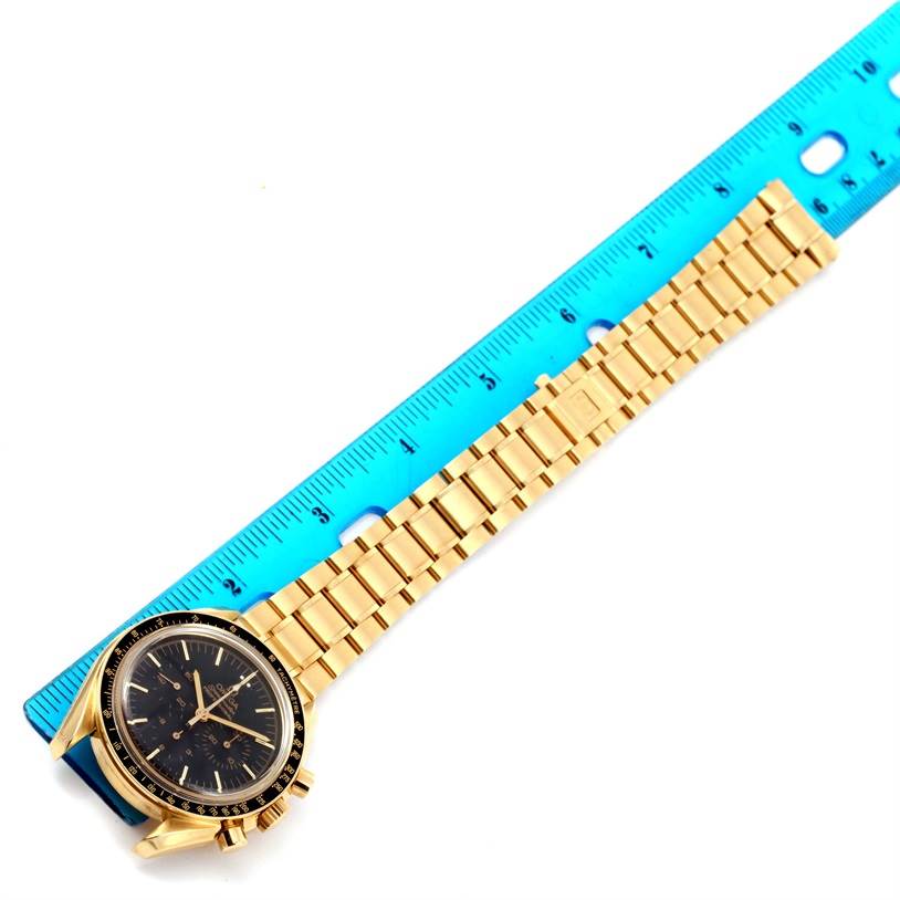 The image shows an Omega Speedmaster 3195.50.00 Men's Yellow Gold Black Dial 3195.50.00 Men's Yellow Gold Black Dial watch laid sideways with its gold bracelet extended alongside a blue ruler.