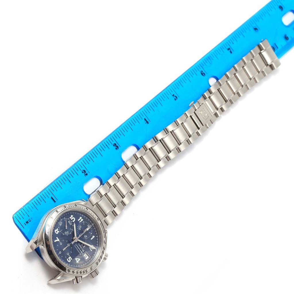 The image shows an Omega Speedmaster 3513.82.00 Men
s Stainless Steel Blue Dial 3513.82.00 Men
s Stainless Steel Blue Dial watch with a stainless steel bracelet laid next to a blue ruler.