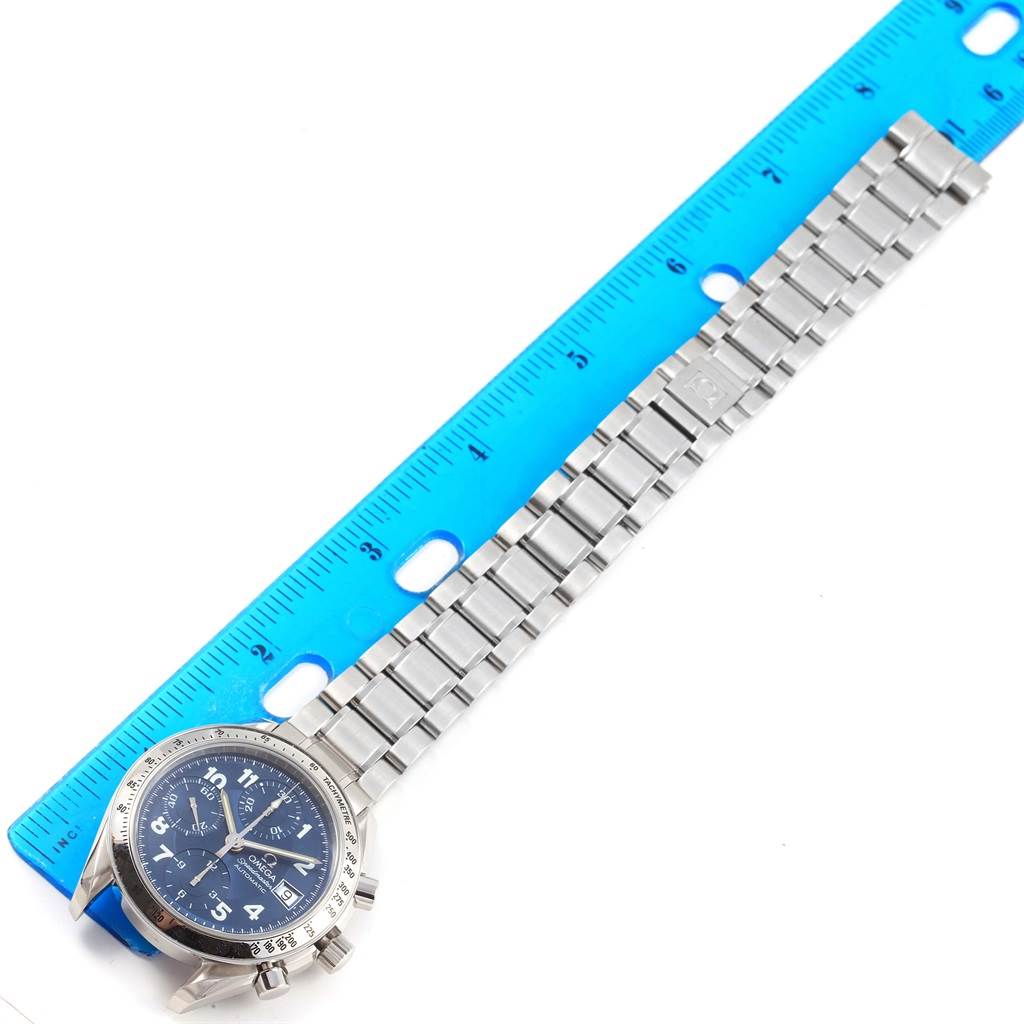 The image shows an Omega Speedmaster 3513.82.00 Men
s Stainless Steel Blue Dial 3513.82.00 Men
s Stainless Steel Blue Dial watch from an overhead angle, with its bracelet placed alongside a blue ruler.