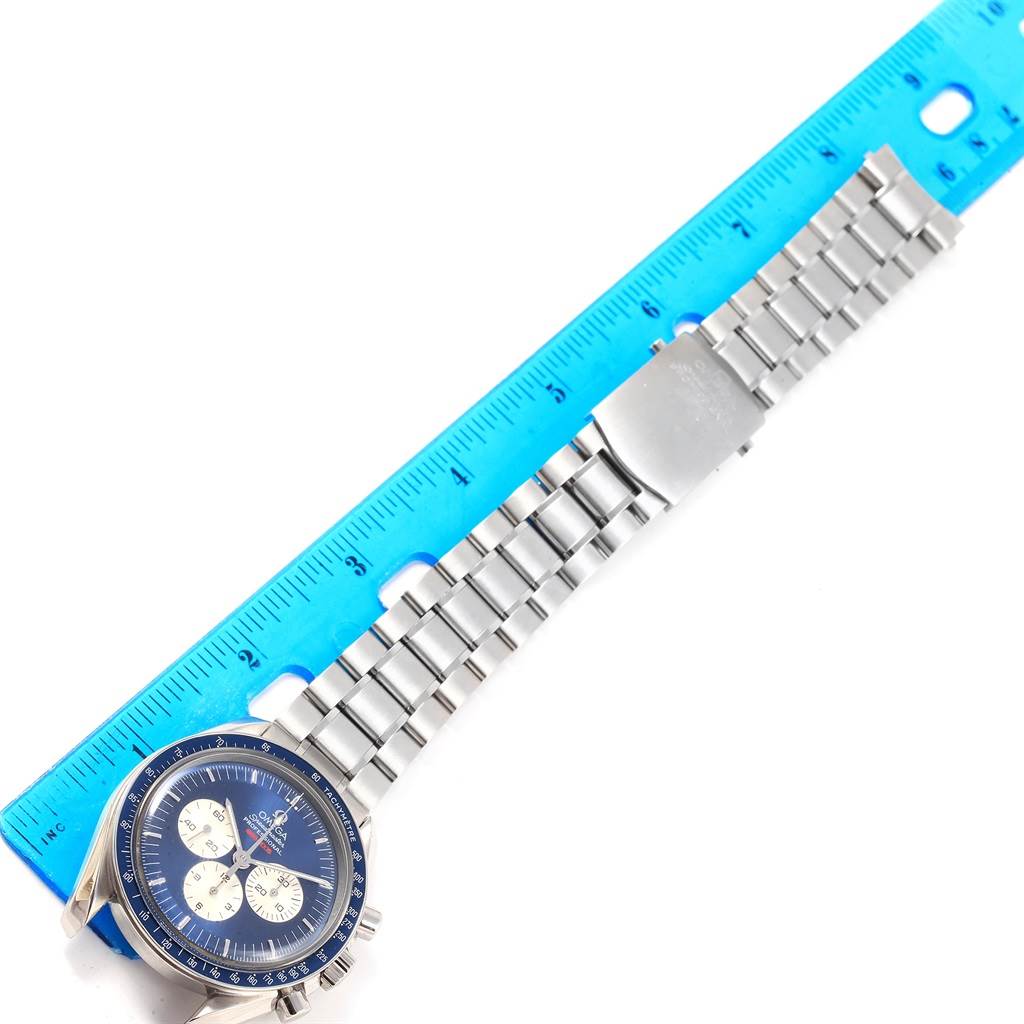 The image shows a top view of an Omega Speedmaster 3565.80.00 Men's Stainless Steel Blue Dial 3565.80.00 Men's Stainless Steel Blue Dial watch with its stainless steel bracelet and clasp, alongside a blue ruler for scale.