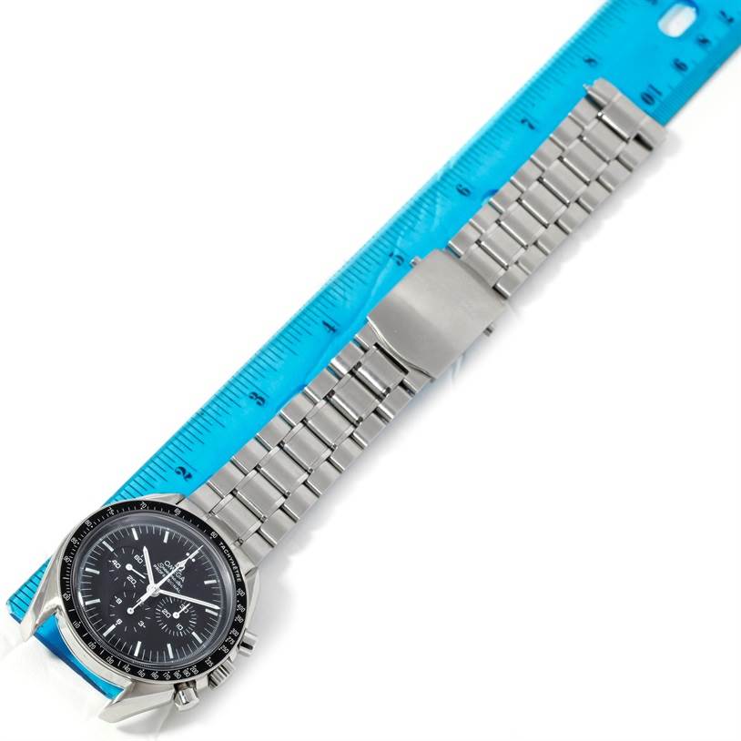 The image shows an Omega Speedmaster Men
s Stainless Steel Black Dial Men
s Stainless Steel Black Dial watch with a stainless steel bracelet lying alongside a blue ruler.