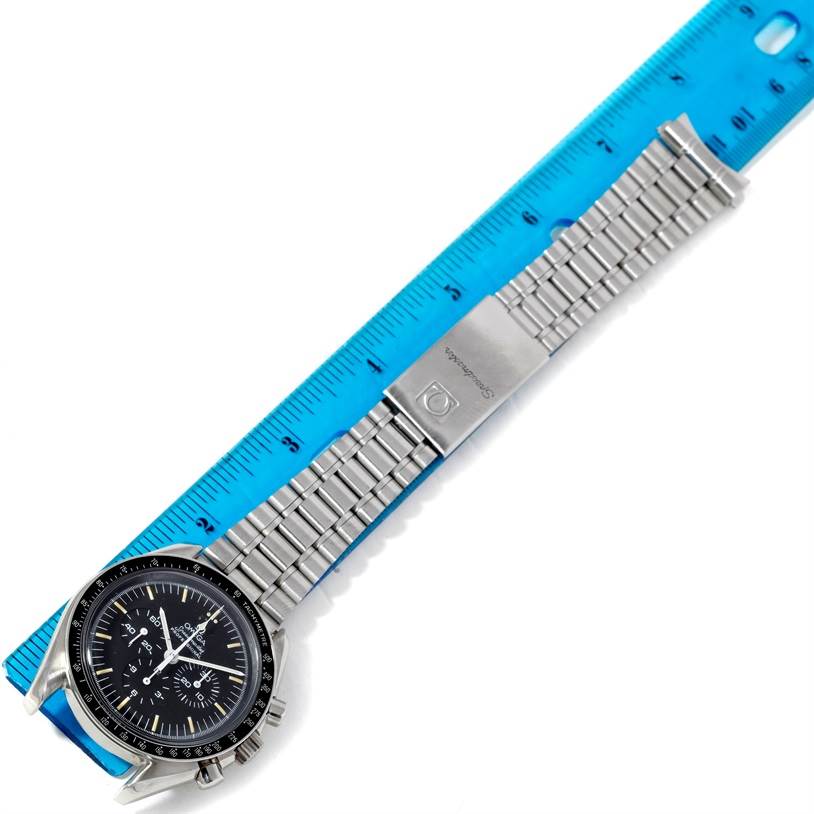 The image shows the Omega Speedmaster Men's Stainless Steel Black Dial Men's Stainless Steel Black Dial watch at a top-down angle, displaying the watch face, stainless steel bracelet, and clasp against a blue ruler.