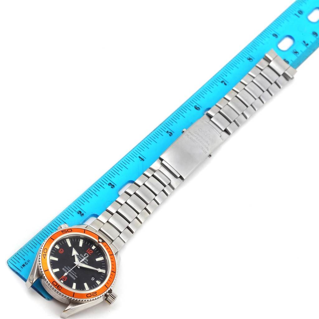 The Omega Planet Ocean 2209.50.00 Men
s Stainless Steel Black Dial 2209.50.00 Men
s Stainless Steel Black Dial watch is displayed flat with its orange bezel, face, stainless steel bracelet, and clasp visible, alongside a blue ruler for scale.