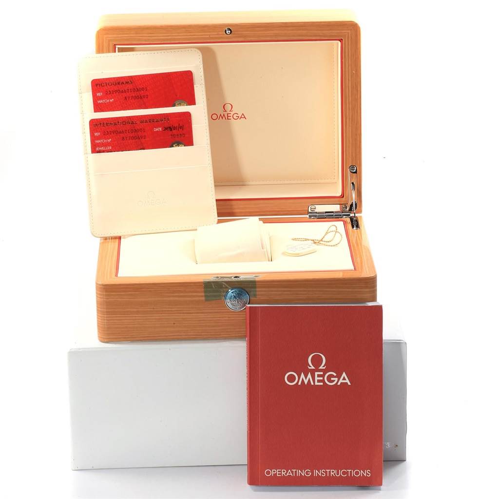 The image shows a wooden box with an Omega logo, warranty cards, and an instruction manual, viewed from a top-front angle.