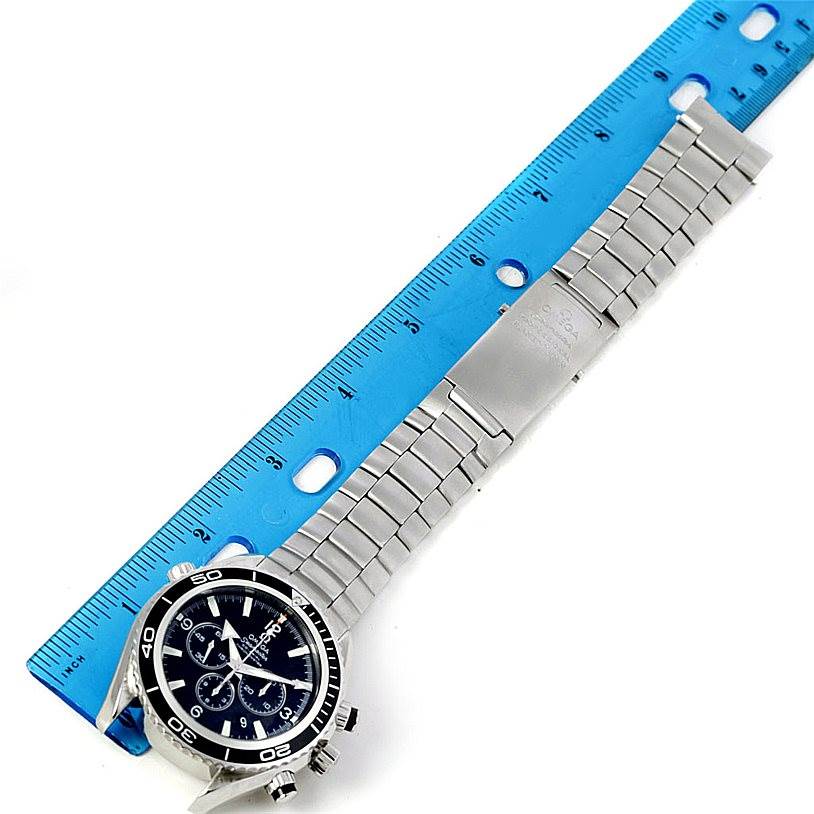 The image shows an Omega Planet Ocean 2210.50.00 Men
s Stainless Steel Black Dial 2210.50.00 Men
s Stainless Steel Black Dial model watch laid flat with a blue ruler next to it, displaying the watch face, bracelet, and clasp.