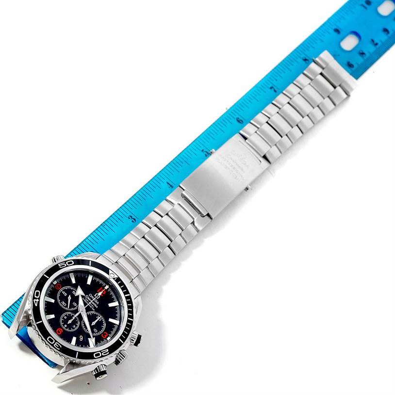 The image shows an Omega Planet Ocean 2210.51.00 Men's Stainless Steel Black Dial 2210.51.00 Men's Stainless Steel Black Dial watch placed next to a blue ruler, highlighting its face, bezel, and metal bracelet.