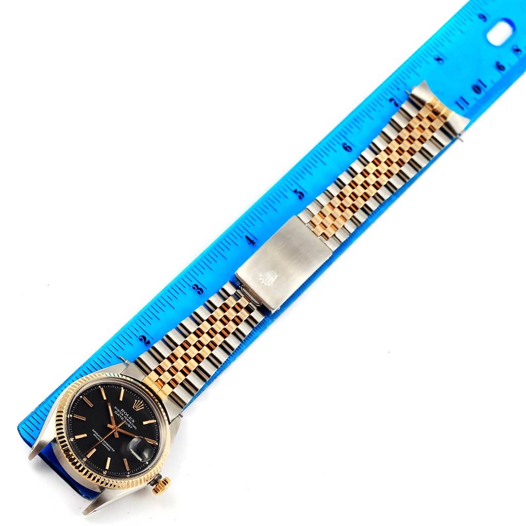 The image shows a Rolex Vintage Collection 1603 Men's Steel and Gold (two tone) Black Dial 1603 Men's Steel and Gold (two tone) Black Dial watch laid flat next to a blue ruler, displaying its face, bezel, bracelet, and clasp.