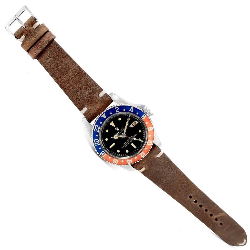 The image shows a top-down view of a Rolex Vintage Collection 1675 Men's Stainless Steel Black Dial 1675 Men's Stainless Steel Black Dial watch with a brown leather strap and a black face with a blue and red bezel.