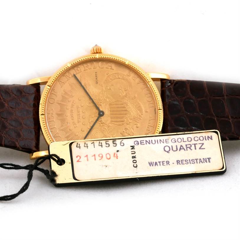 The image shows a frontal view of the Corum Coin Watch Men's Yellow Gold Black Dial Men's Yellow Gold Black Dial model, showcasing its gold coin face with a brown leather strap and attached tag.