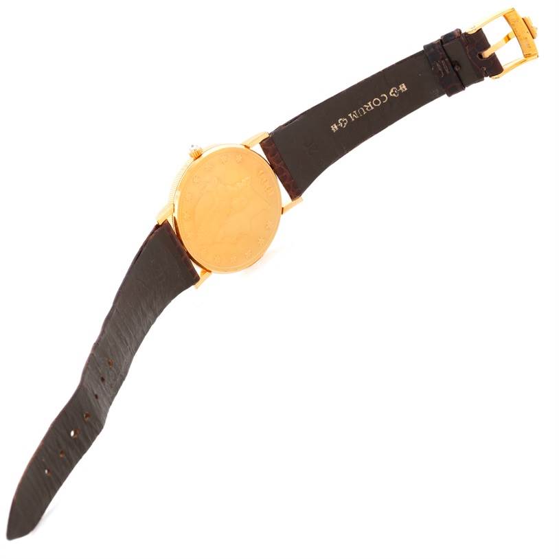 The Coin Watch model by Corum is shown from a top angle, displaying the case back, leather strap, and gold buckle.