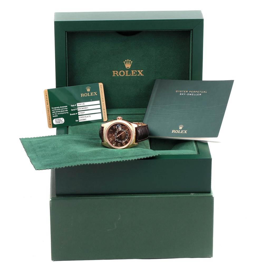 The image shows the Rolex Sky-Dweller 326135 Men's Rose Gold Brown Dial 326135 Men's Rose Gold Brown Dial watch with its box, warranty card, manual, and cleaning cloth.