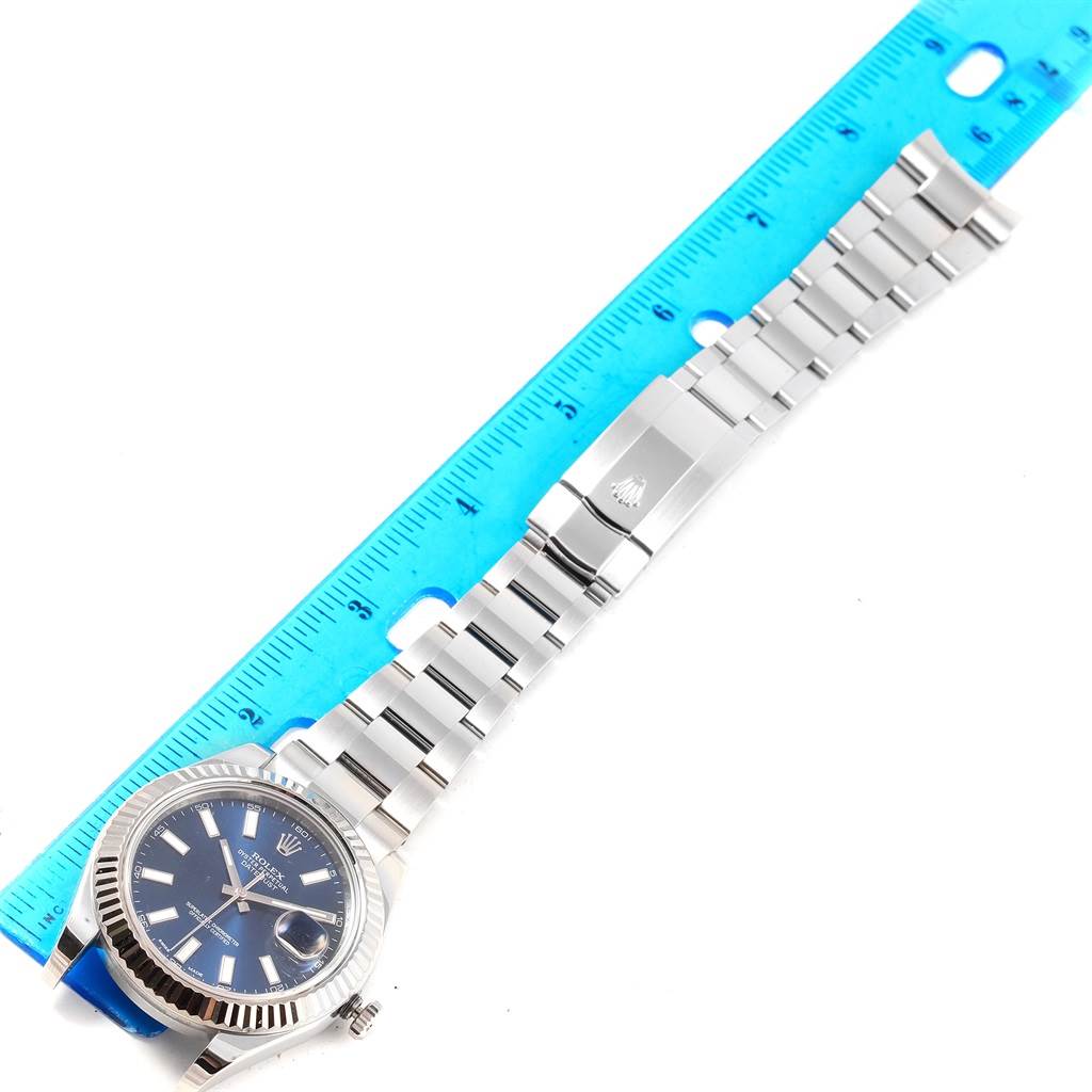 The image shows a top-down view of a Rolex Datejust 41 116334 Men's Stainless Steel Blue Dial 116334 Men's Stainless Steel Blue Dial model watch with its blue dial and metallic bracelet placed next to a blue ruler.