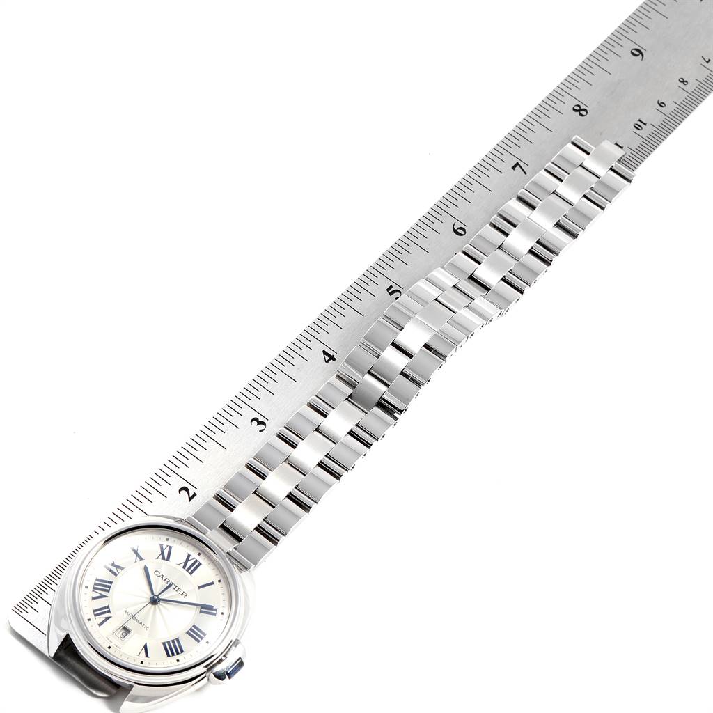The image shows a Cle model by Cartier watch laid next to a ruler, highlighting the watch face and bracelet.