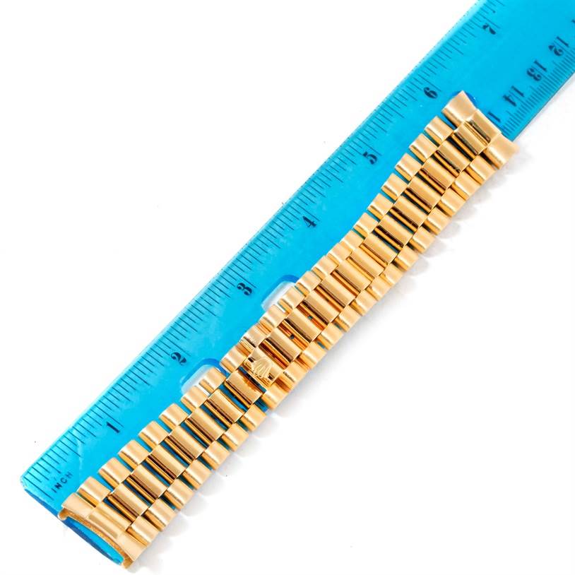 This image shows the gold bracelet of a Rolex President   model watch next to a blue ruler, viewed from above.