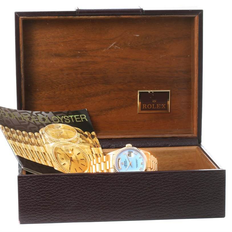 The Rolex President 18038 Men
s Yellow Gold Mother of Pearl Dial 18038 Men
s Yellow Gold Mother of Pearl Dial model watch is displayed at an angle, partially inside an open Rolex-branded wooden box, alongside booklets.