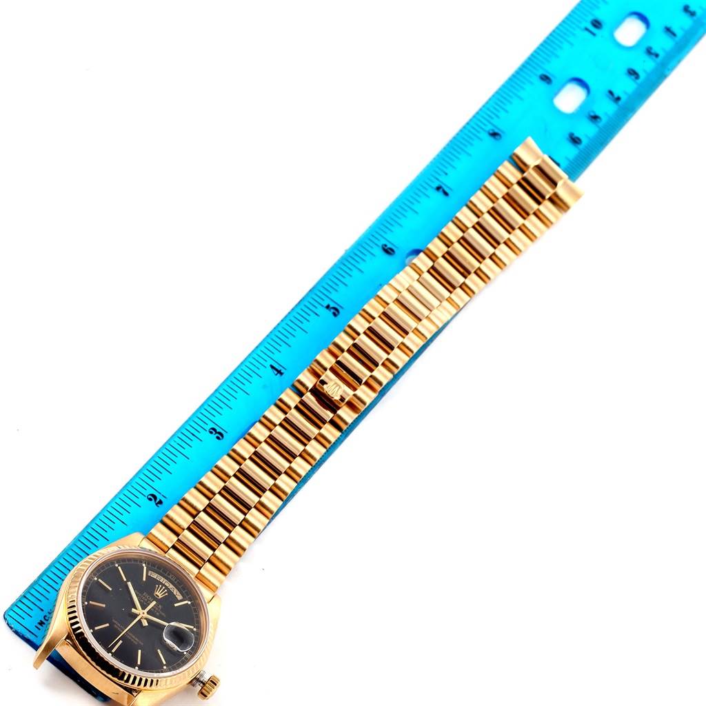 The Rolex President 18038 Men
s Yellow Gold Black Dial 18038 Men
s Yellow Gold Black Dial model watch is shown from a top view, featuring the watch face and linked bracelet next to a ruler.