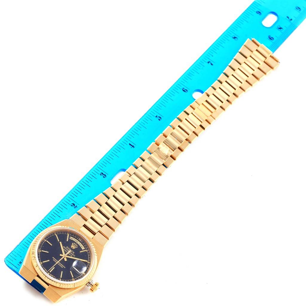 The image shows a Rolex President 19018 Men
s Yellow Gold Black Dial 19018 Men
s Yellow Gold Black Dial model watch placed next to a blue ruler, displaying the face and entire bracelet.