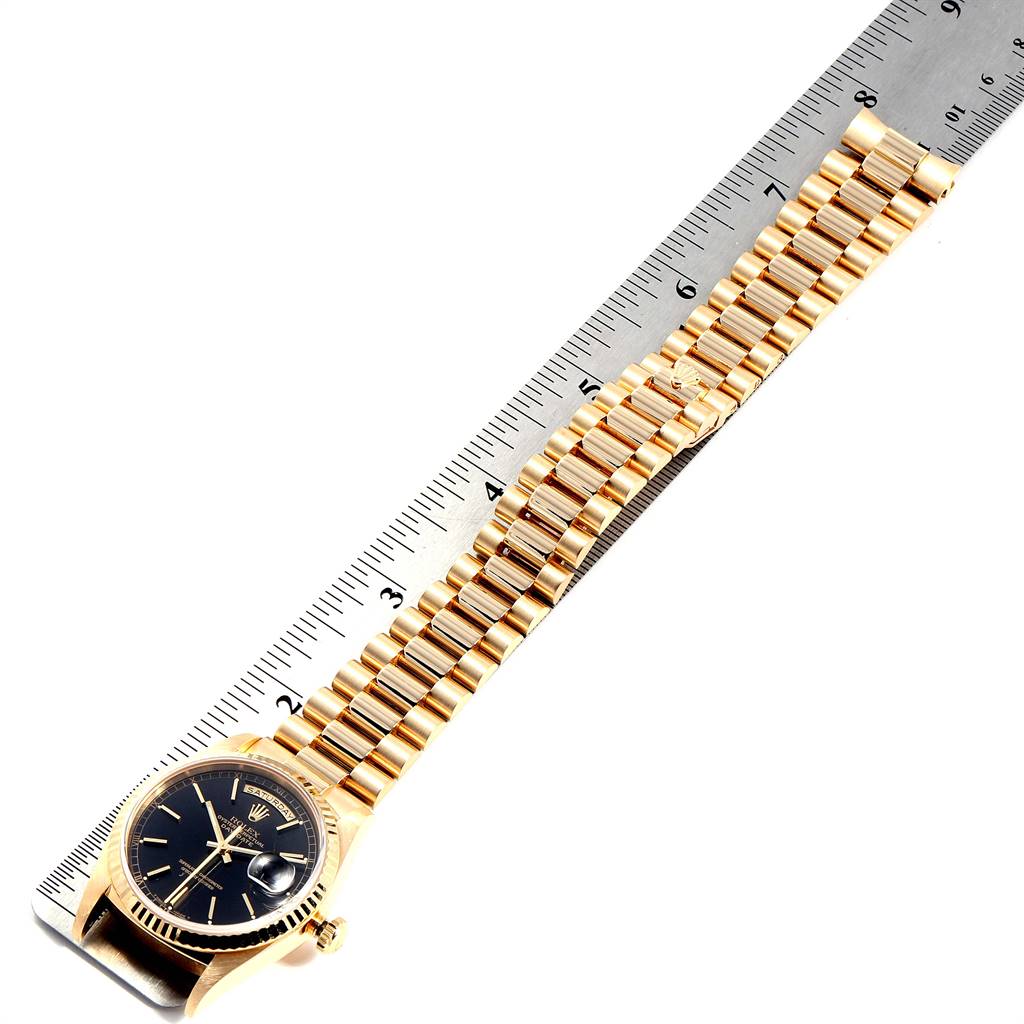 This Rolex President 18238 Men
s Yellow Gold Black Dial 18238 Men
s Yellow Gold Black Dial model watch is shown at an angle displaying the entire bracelet and face, laid next to a ruler.