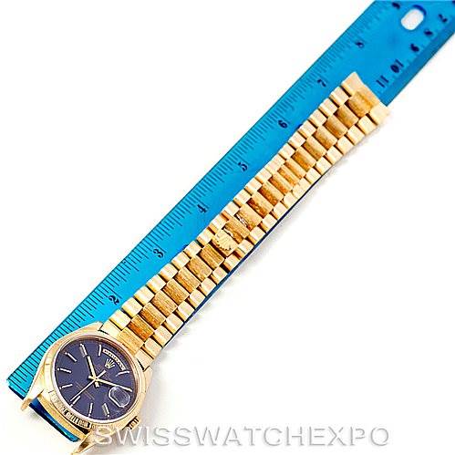 The image shows a horizontal view of a Rolex President 18248 Men's Yellow Gold Blue Dial 18248 Men's Yellow Gold Blue Dial model watch with a blue dial and gold bracelet placed alongside a blue ruler.