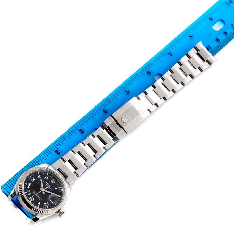 The Rolex Datejust 115234 Men
s Stainless Steel Black Dial 115234 Men
s Stainless Steel Black Dial watch is displayed from a top-down angle, with the full band extended next to a blue ruler.