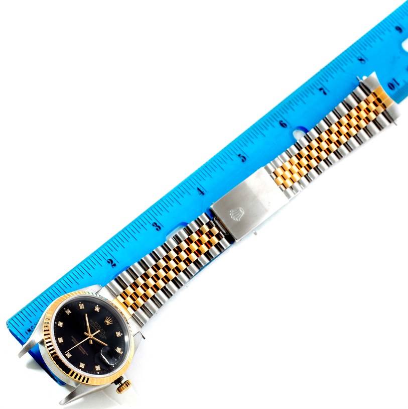 This image shows a Rolex Datejust 16233 Men's Steel and Gold (two tone) Black Dial 16233 Men's Steel and Gold (two tone) Black Dial watch lying flat with its two-tone band next to a blue measuring ruler.