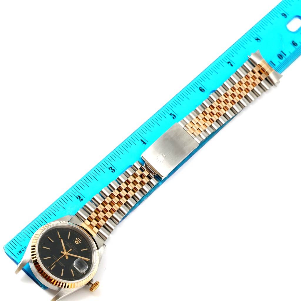 The image shows a Rolex Datejust 16233 Men's Steel and Gold (two tone) Black Dial 16233 Men's Steel and Gold (two tone) Black Dial watch laid flat with its band extended, alongside a blue ruler for scale.
