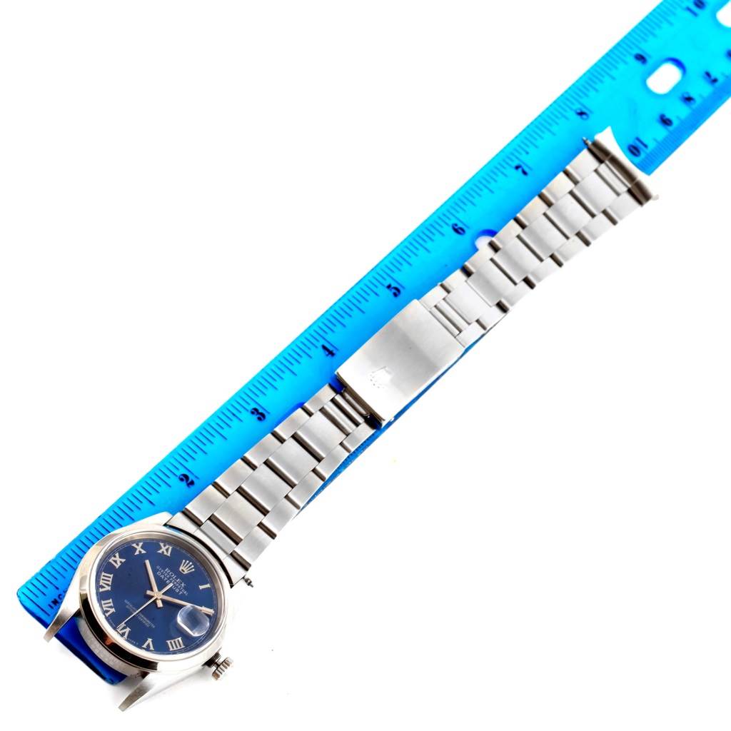 The image shows a Rolex Datejust 16200 Men's Stainless Steel Blue Dial 16200 Men's Stainless Steel Blue Dial watch laid horizontally next to a blue ruler, displaying its face, bracelet, and clasp.