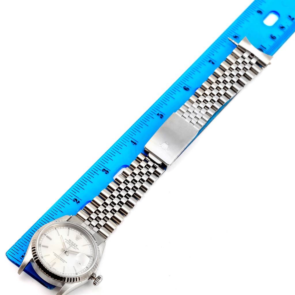 The Rolex Datejust 16234 Men
s Stainless Steel Silver Dial 16234 Men
s Stainless Steel Silver Dial watch is shown laid flat next to a blue ruler, displaying its face, case, and Jubilee bracelet band.