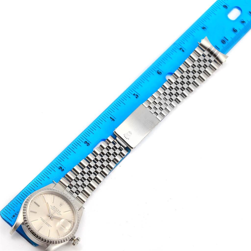 The image shows a top view of a Rolex Datejust 16220 Men
s Stainless Steel Silver Dial 16220 Men
s Stainless Steel Silver Dial watch with its band extended alongside a blue ruler.