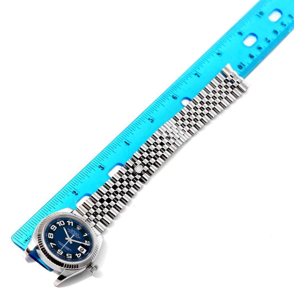 The image shows a Rolex Datejust 116234 Men
s Stainless Steel Blue Dial 116234 Men
s Stainless Steel Blue Dial watch laid next to a blue ruler, highlighting its face and metal bracelet in a side view.