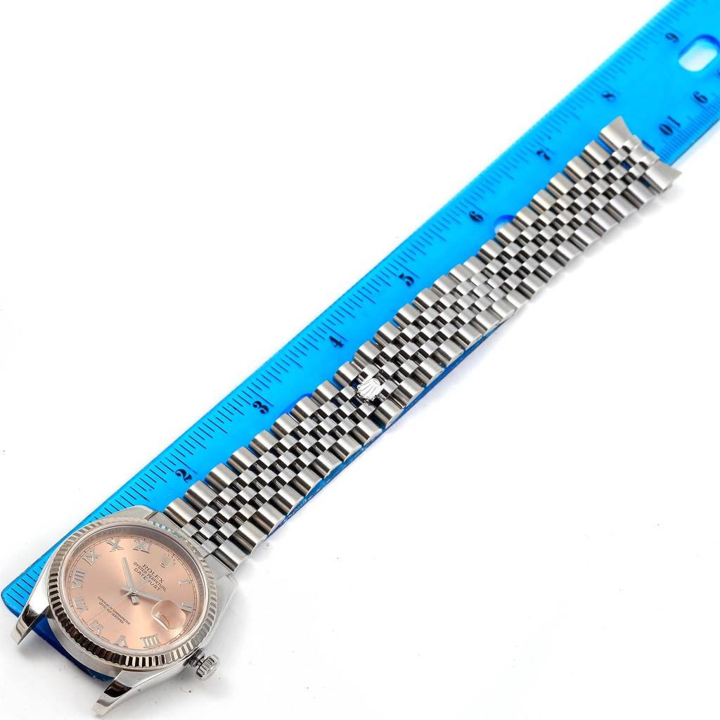 The Rolex Datejust 116234 Stainless Steel Pink Dial 116234 Stainless Steel Pink Dial watch is shown from a top-down angle, displaying its face and fully extended metal bracelet next to a blue ruler.