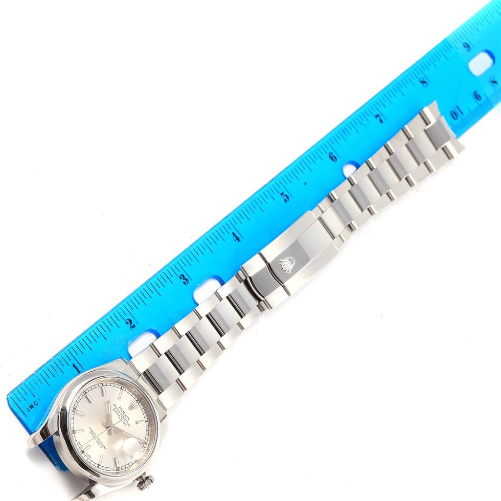 This image shows the Rolex Datejust 116200 Men's Stainless Steel Silver Dial 116200 Men's Stainless Steel Silver Dial watch lying flat with its band extended and measured next to a blue ruler.