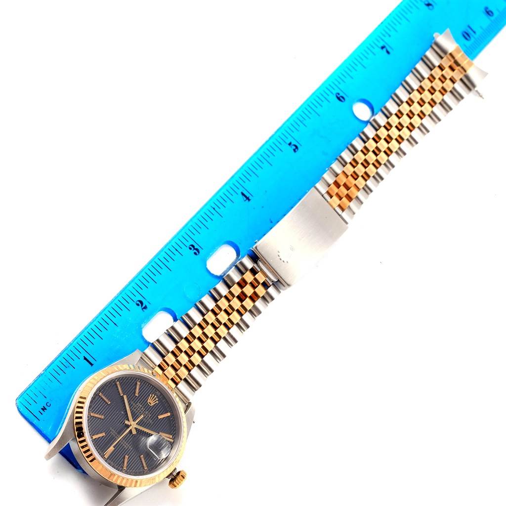 The image shows the Rolex Datejust 16233 Men's Steel and Gold (two tone) Black Dial 16233 Men's Steel and Gold (two tone) Black Dial watch face and part of the band next to a blue ruler for scale.