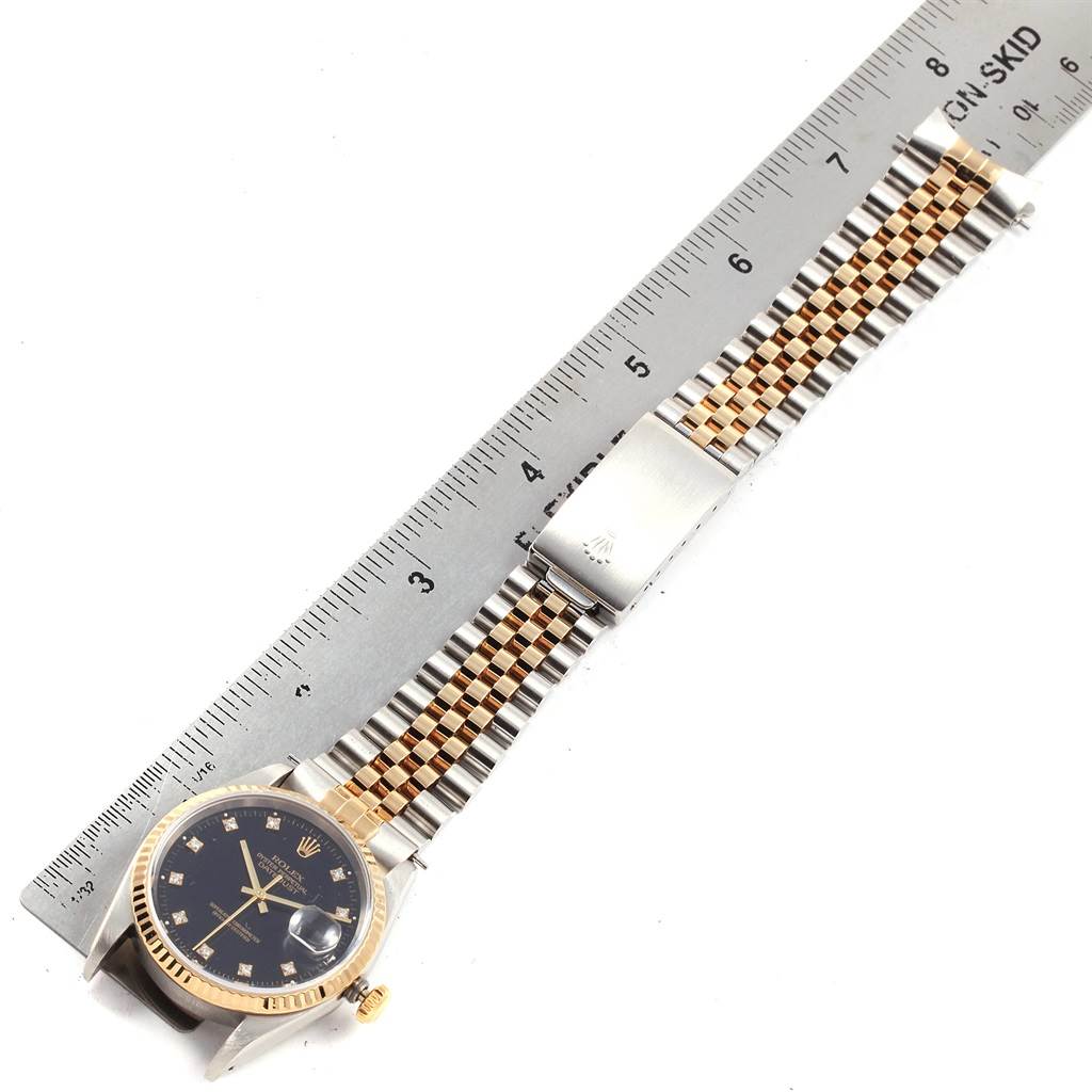The image shows a Rolex Datejust 16233 Men's Steel and Gold (two tone) Black Dial 16233 Men's Steel and Gold (two tone) Black Dial watch laid flat next to a ruler, highlighting its band, clasp, and face.