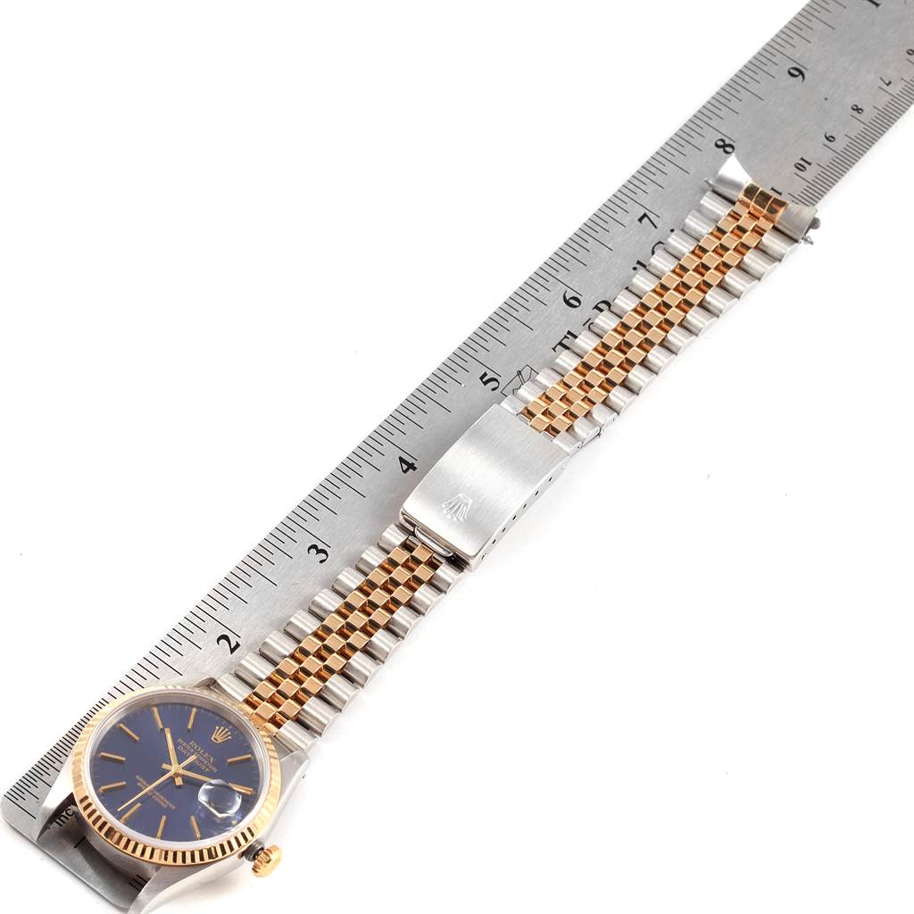 The Rolex Datejust 16233 Men
s Steel and Gold (two tone) Blue Dial 16233 Men
s Steel and Gold (two tone) Blue Dial watch is shown in a top-down angle, showcasing its face, Jubilee bracelet, and clasp alongside a ruler.