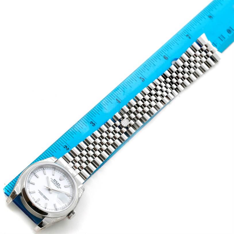 The image shows a Rolex Datejust 116200 Men
s Stainless Steel White Dial 116200 Men
s Stainless Steel White Dial watch with a silver dial and Jubilee bracelet beside a blue ruler for measurement.