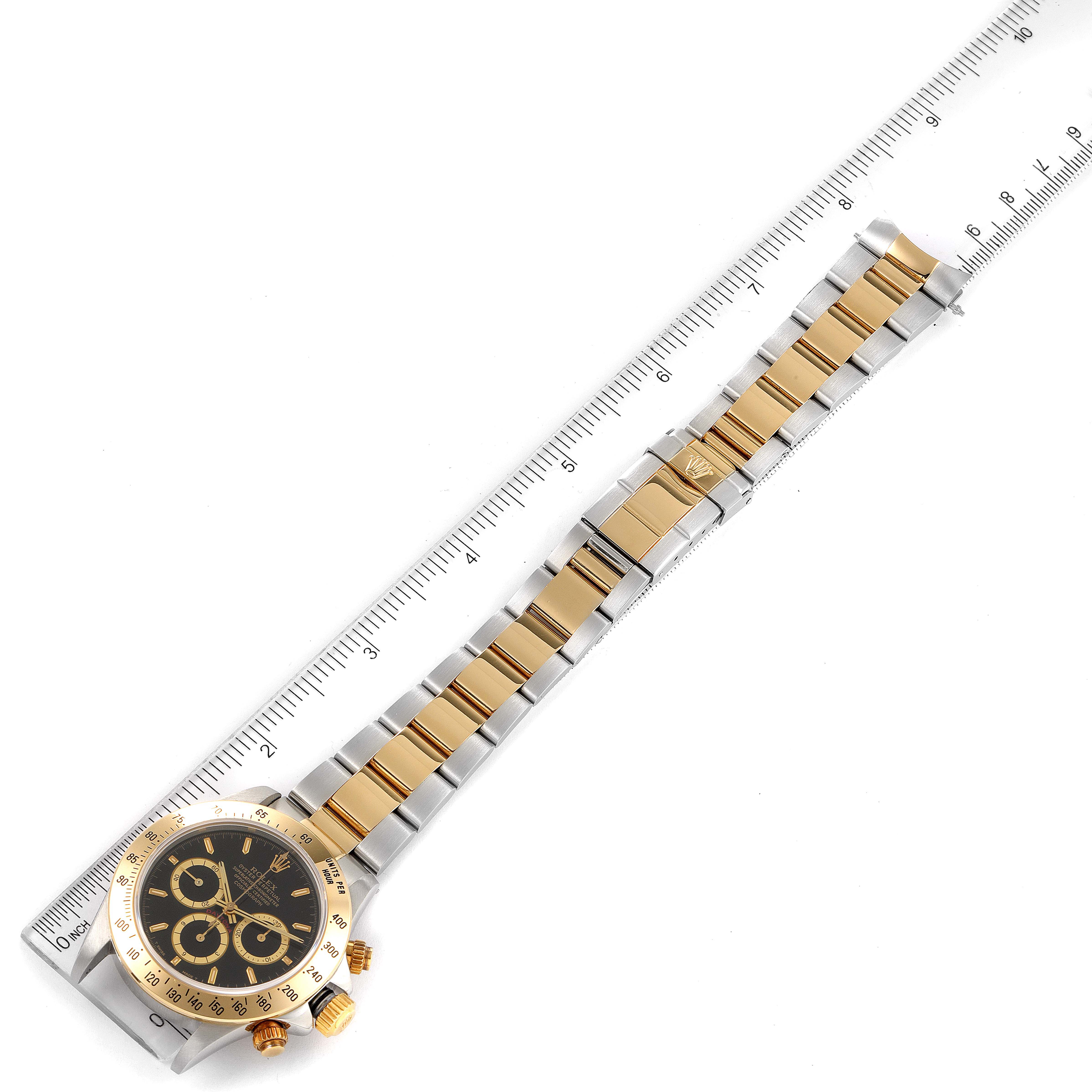 The image shows a Rolex Daytona 16523 Men
s Steel and Gold (two tone) Black Dial 16523 Men
s Steel and Gold (two tone) Black Dial watch laid flat with the entire bracelet and face visible next to a ruler for scale.