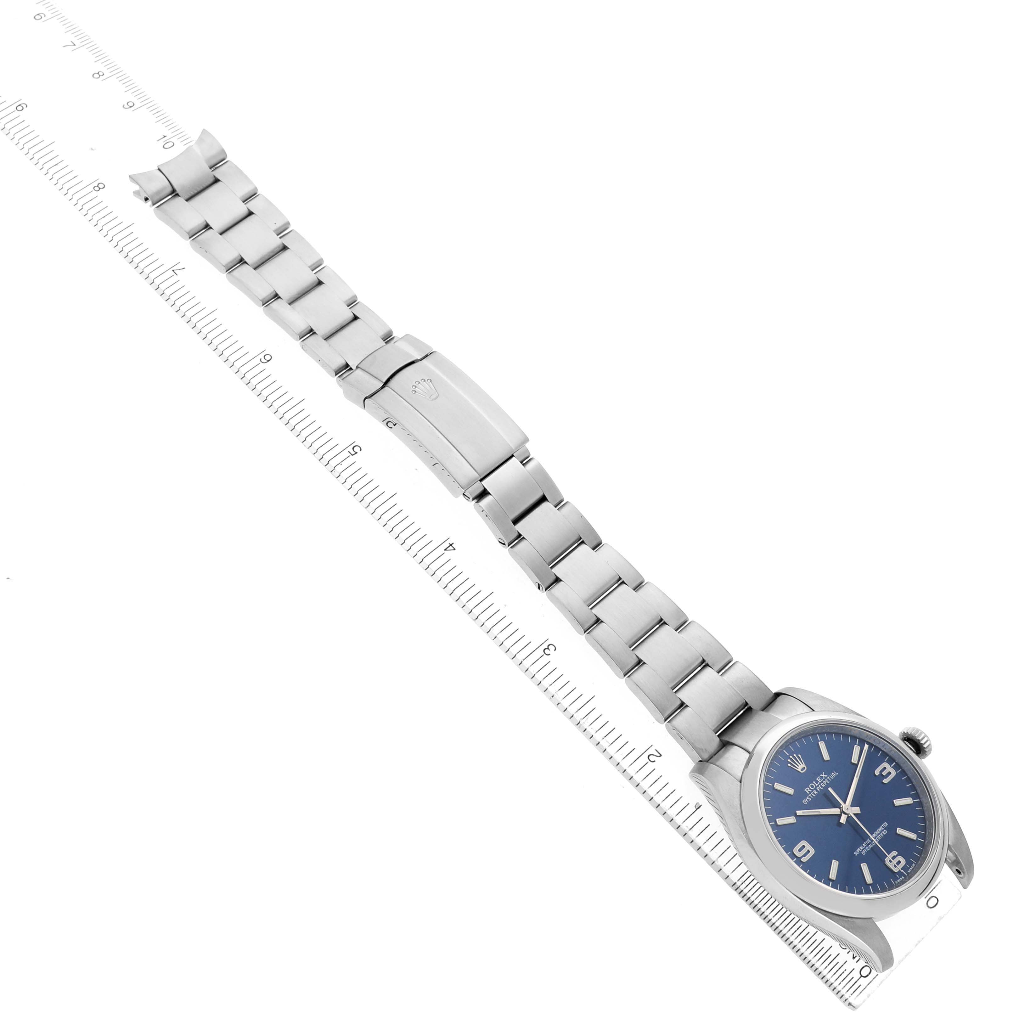 The Rolex Oyster Perpetual 116000 Men's Stainless Steel Blue Dial watch is shown diagonally displaying the face, bracelet, and clasp along a ruler.