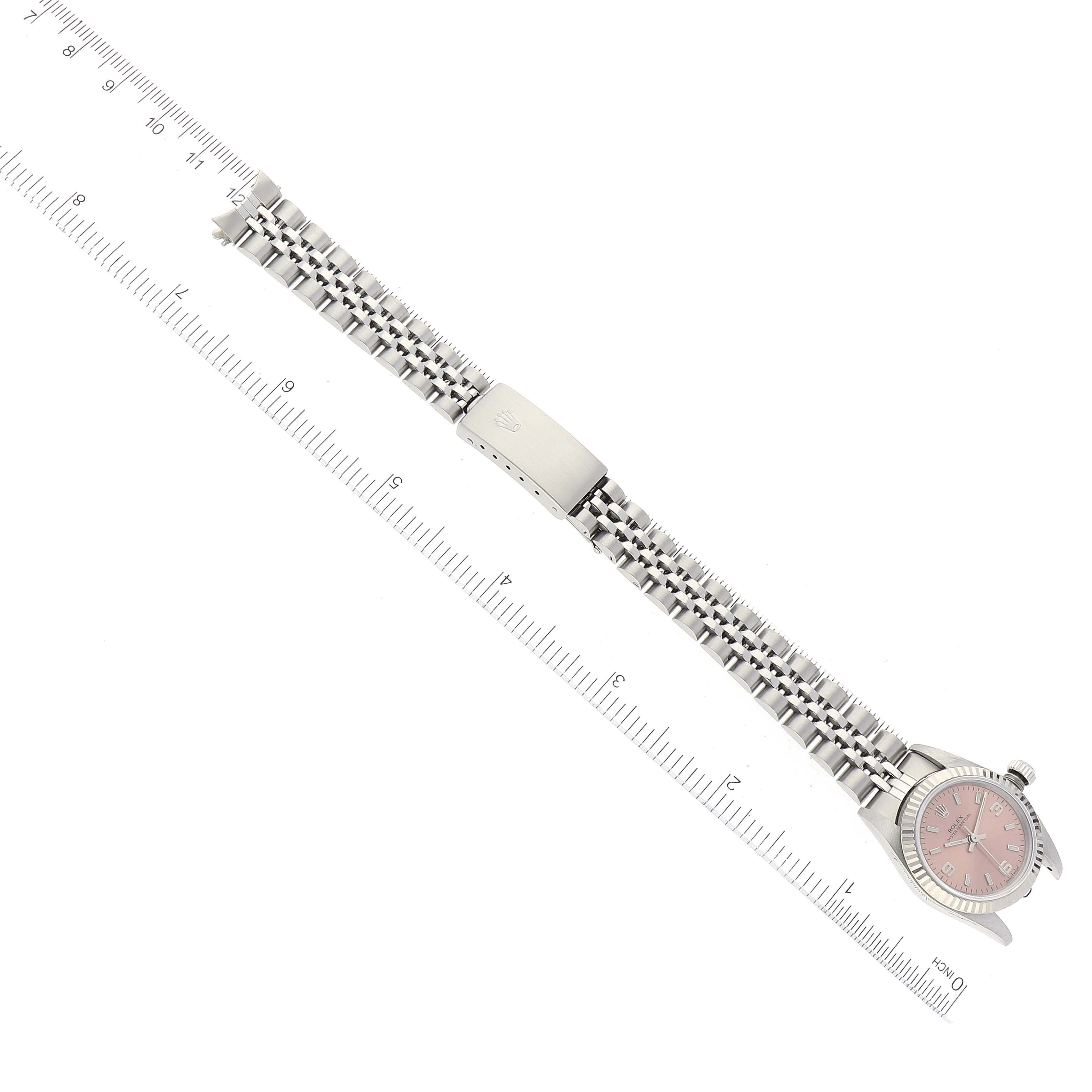 The image shows a Rolex Oyster Perpetual 76094 Women
s Stainless Steel Pink Dial 76094 Women
s Stainless Steel Pink Dial watch lying flat, displaying its metal bracelet and clasp, alongside measuring rulers.