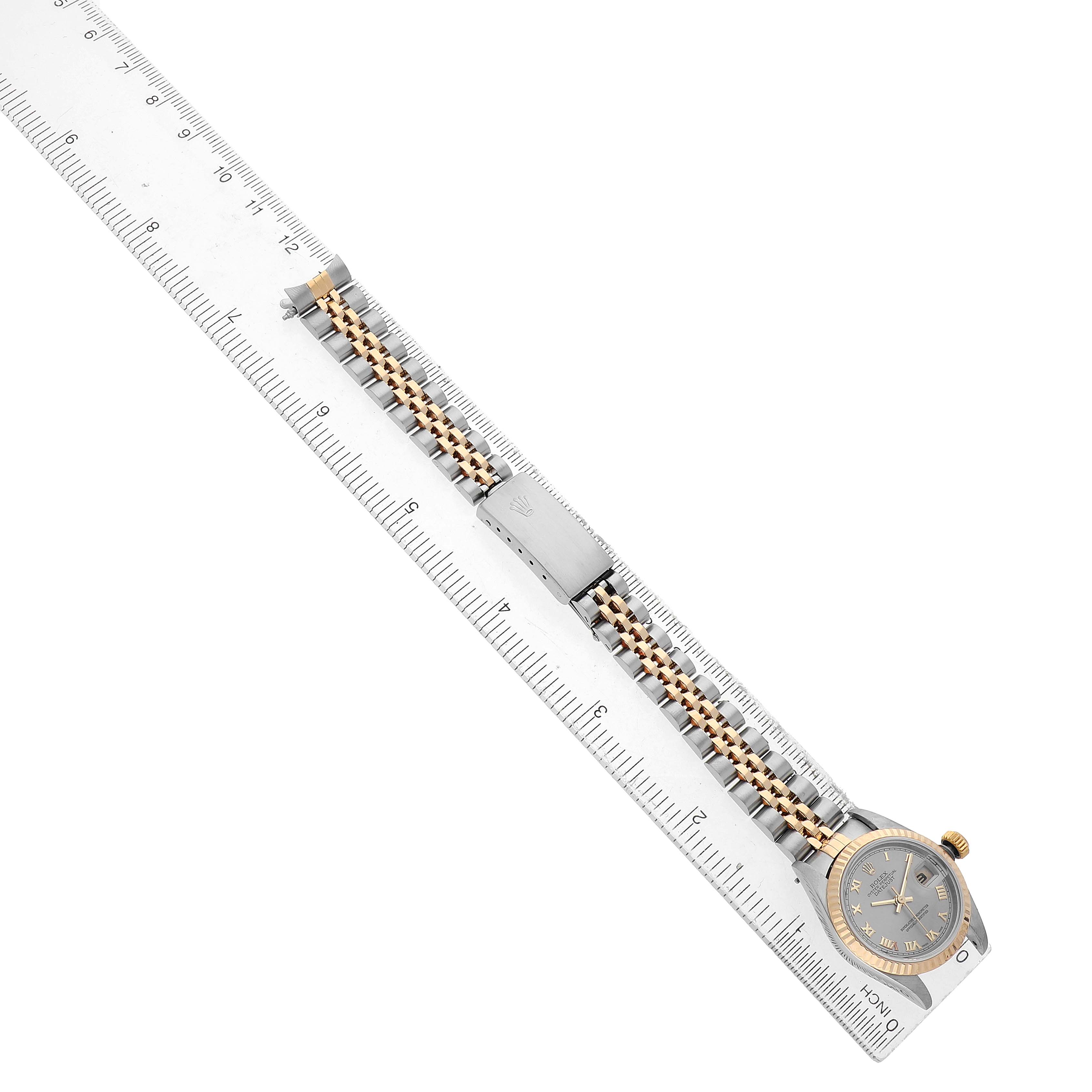 The Rolex Datejust 79173 Women's Steel and Gold (two tone) Silver Dial watch is shown from a top view, displaying the dial, bezel, and bracelet on a ruler.