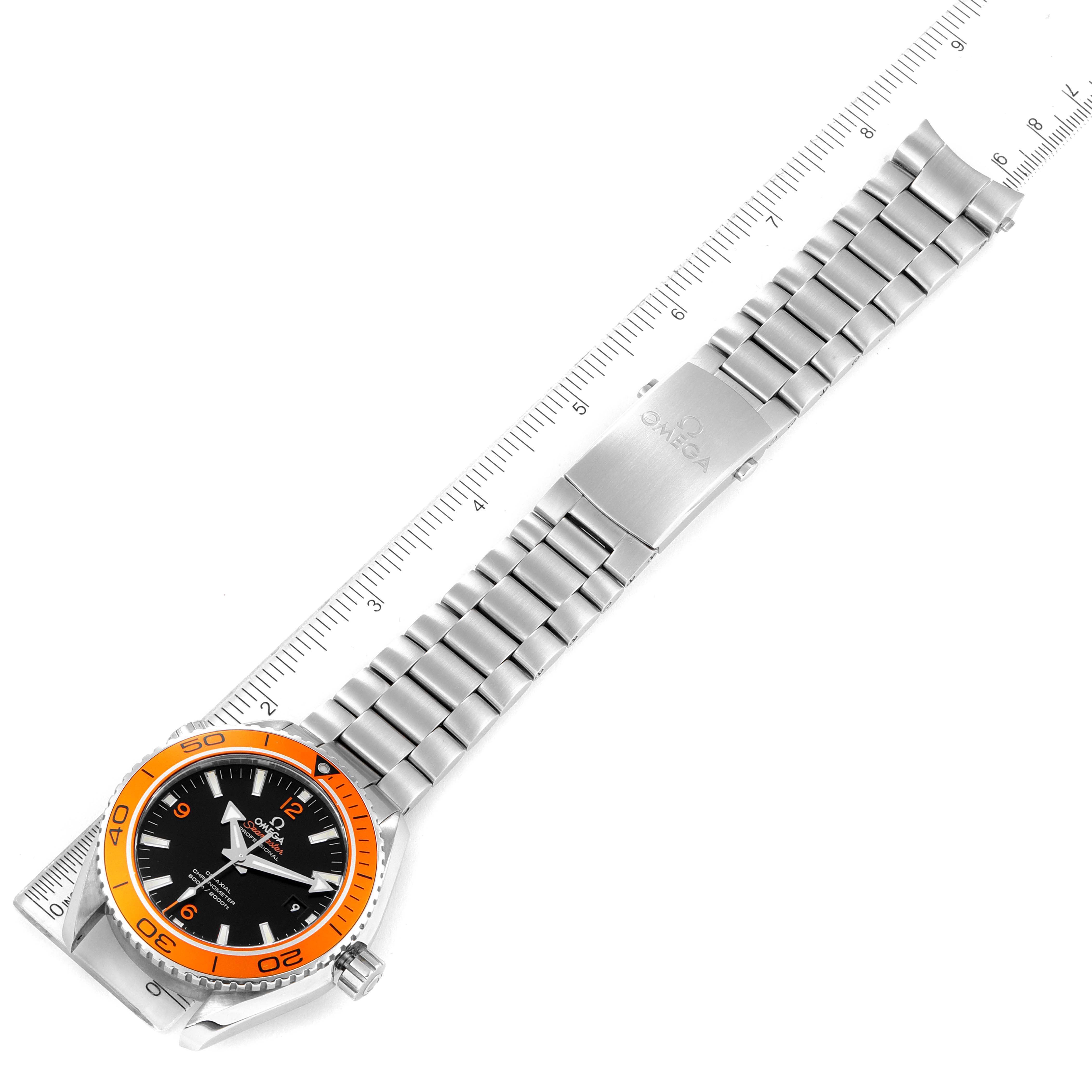 The image shows an Omega Planet Ocean 232.30.46.21.01.002 Men's Stainless Steel Black Dial 232.30.46.21.01.002 Men's Stainless Steel Black Dial watch from a top-down angle, displaying its orange bezel, black dial, and metallic bracelet next to a ruler.