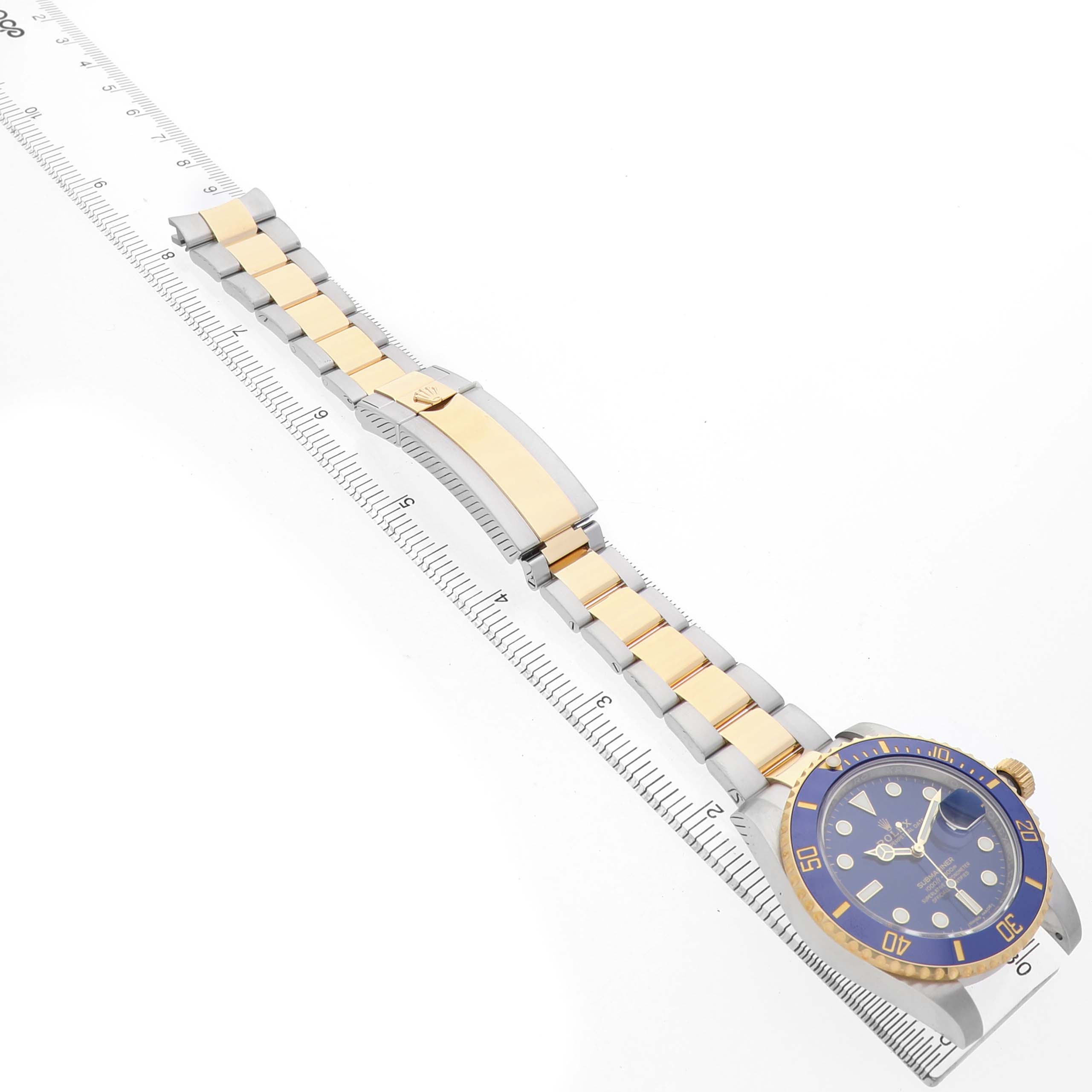 The Rolex Submariner 116613 Men's Steel and Gold (two tone) Blue Dial watch is angled diagonally, showing the face, bezel, bracelet, and clasp on a ruler.