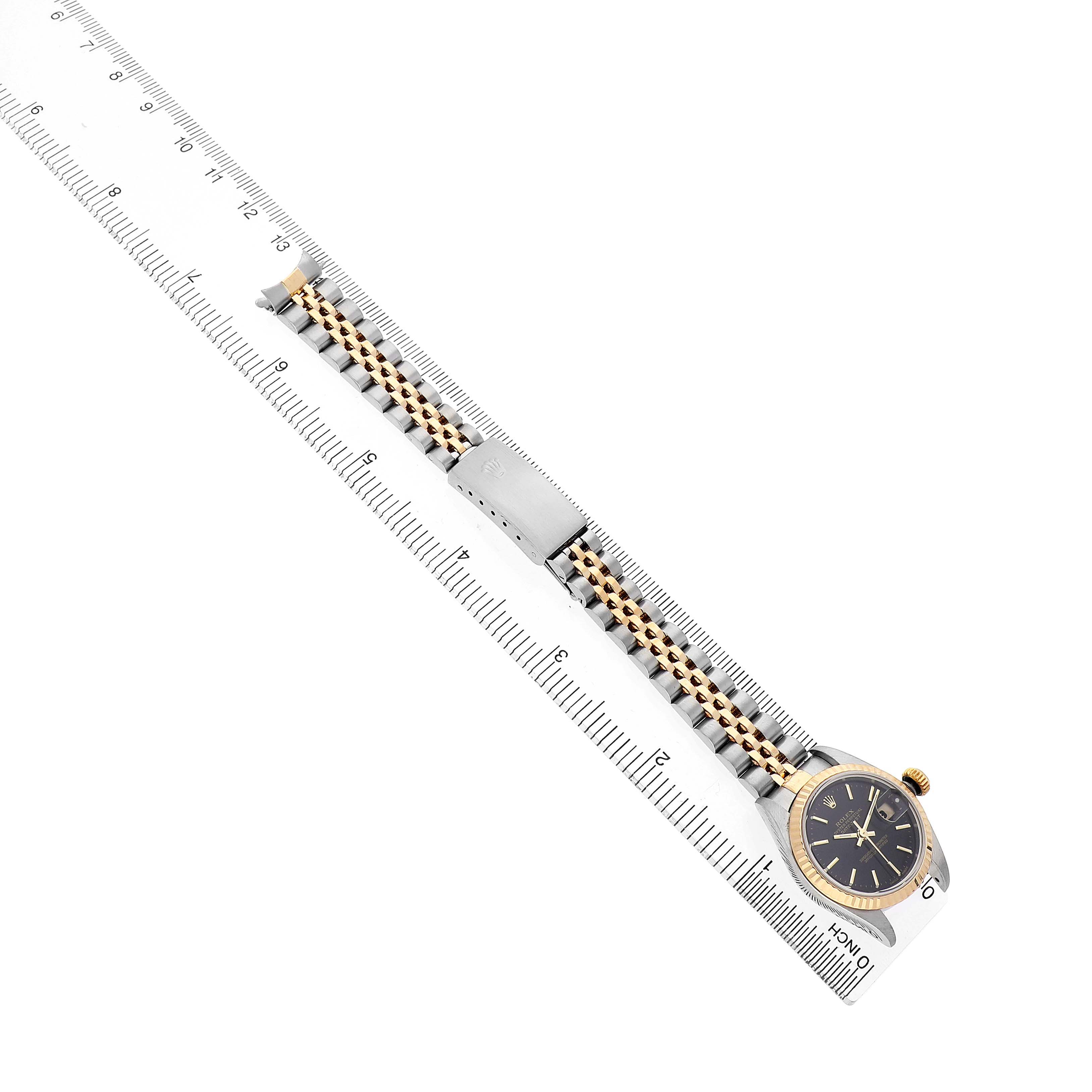 The Rolex Datejust 79173 Women's Steel and Gold (two tone) Black Dial watch is shown diagonally, displaying the face, bezel, and part of the bracelet against a ruler for scale.