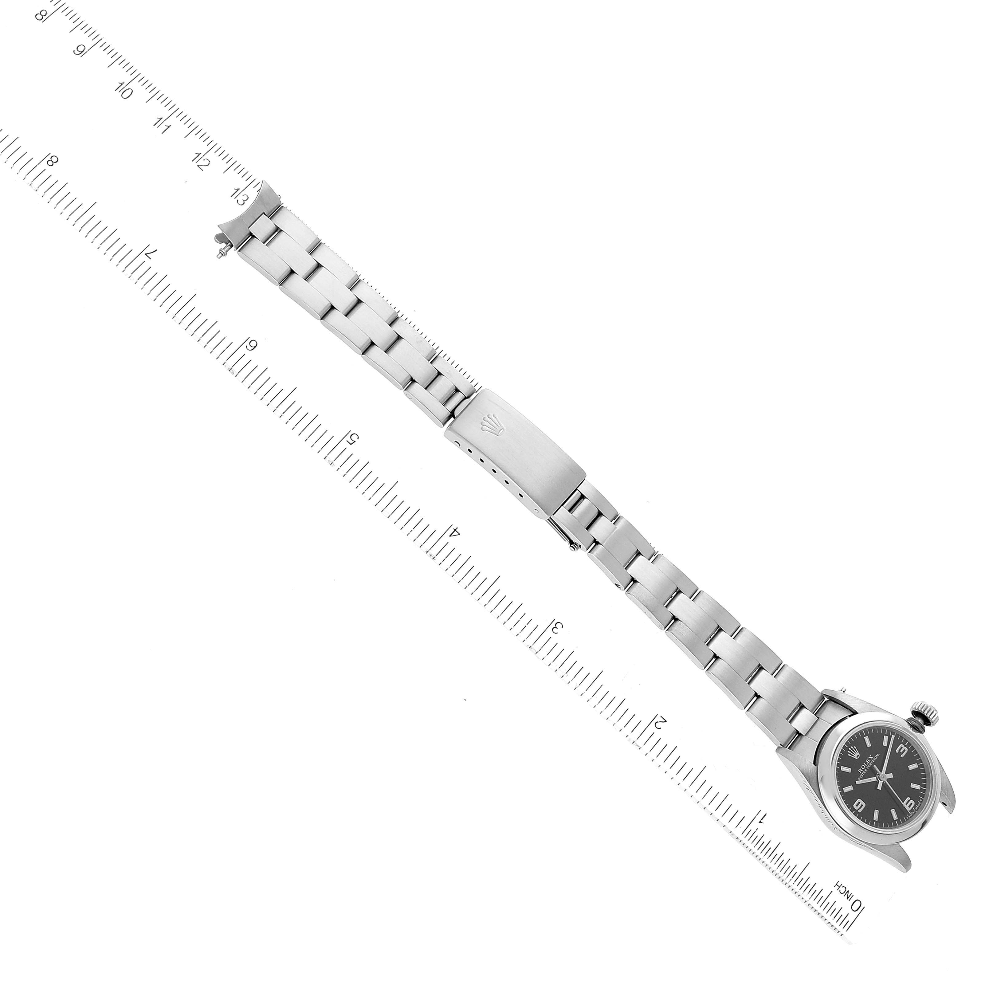 The image shows a top-down view of a Rolex Oyster Perpetual 67180 Women's Stainless Steel Black Dial 67180 Women's Stainless Steel Black Dial watch, highlighting its face, band, and clasp with a ruler for scale.