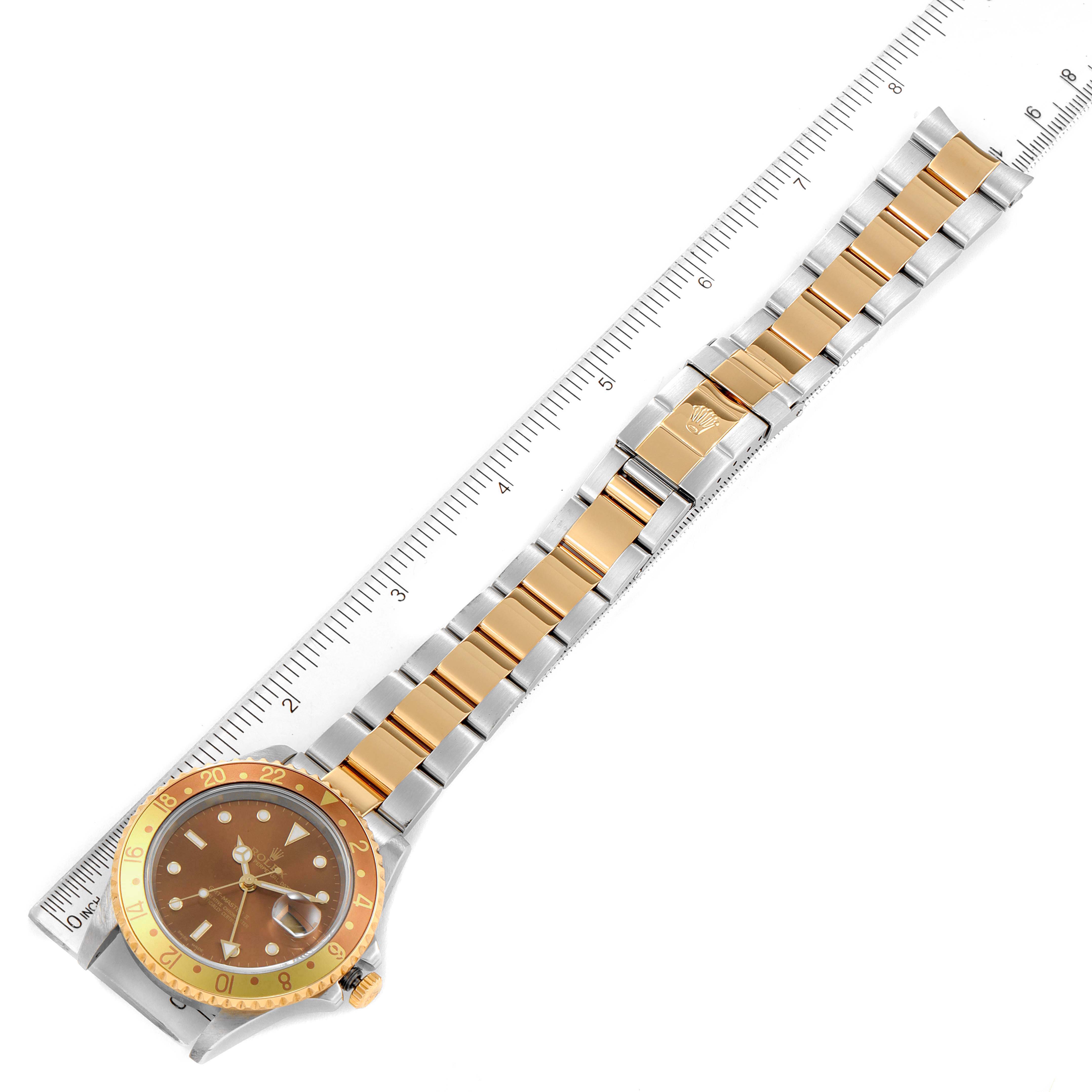 This image shows a Rolex GMT-Master 16713 Men's Steel and Gold (two tone) Brown Dial 16713 Men's Steel and Gold (two tone) Brown Dial watch laid flat, showcasing its dial, bezel, and bracelet with a ruler for scale.