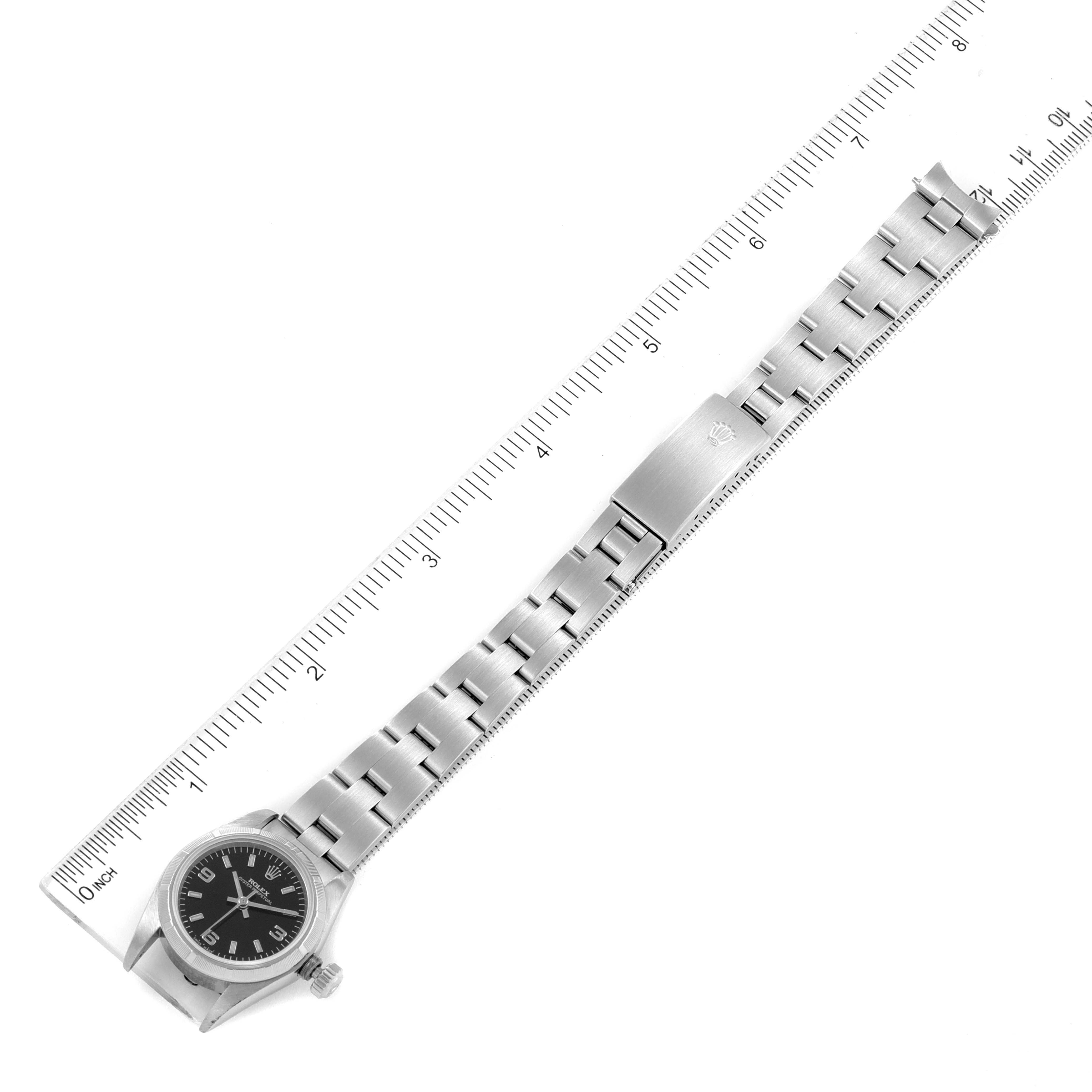The image shows a top view of a Rolex Oyster Perpetual 67230 Women's Stainless Steel Black Dial 67230 Women's Stainless Steel Black Dial watch, including the watch face, bracelet, and clasp, with a ruler for scale.
