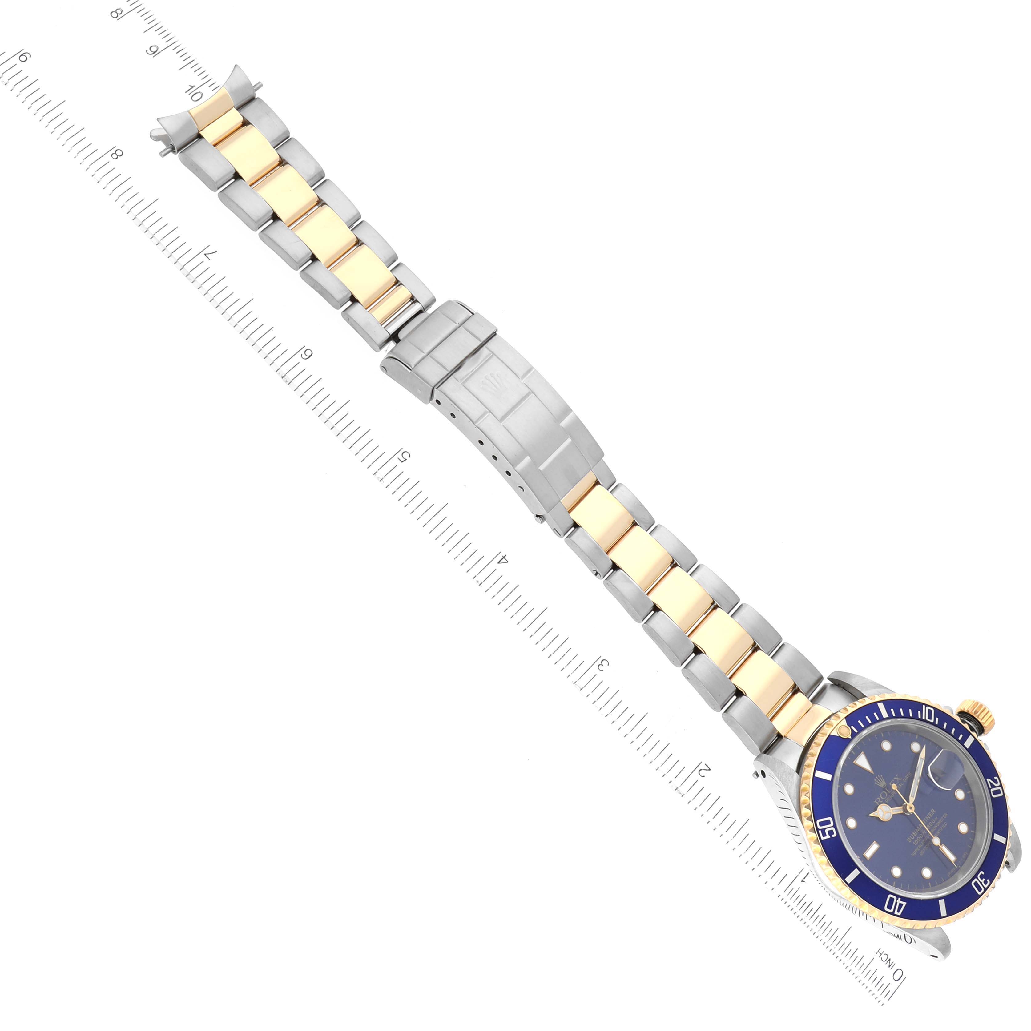 The image shows a Rolex Submariner 16613 Men's Steel and Gold (two tone) Blue Dial 16613 Men's Steel and Gold (two tone) Blue Dial watch laid flat, displaying its blue dial, two-tone metal bracelet, and clasp, with a ruler for scale.