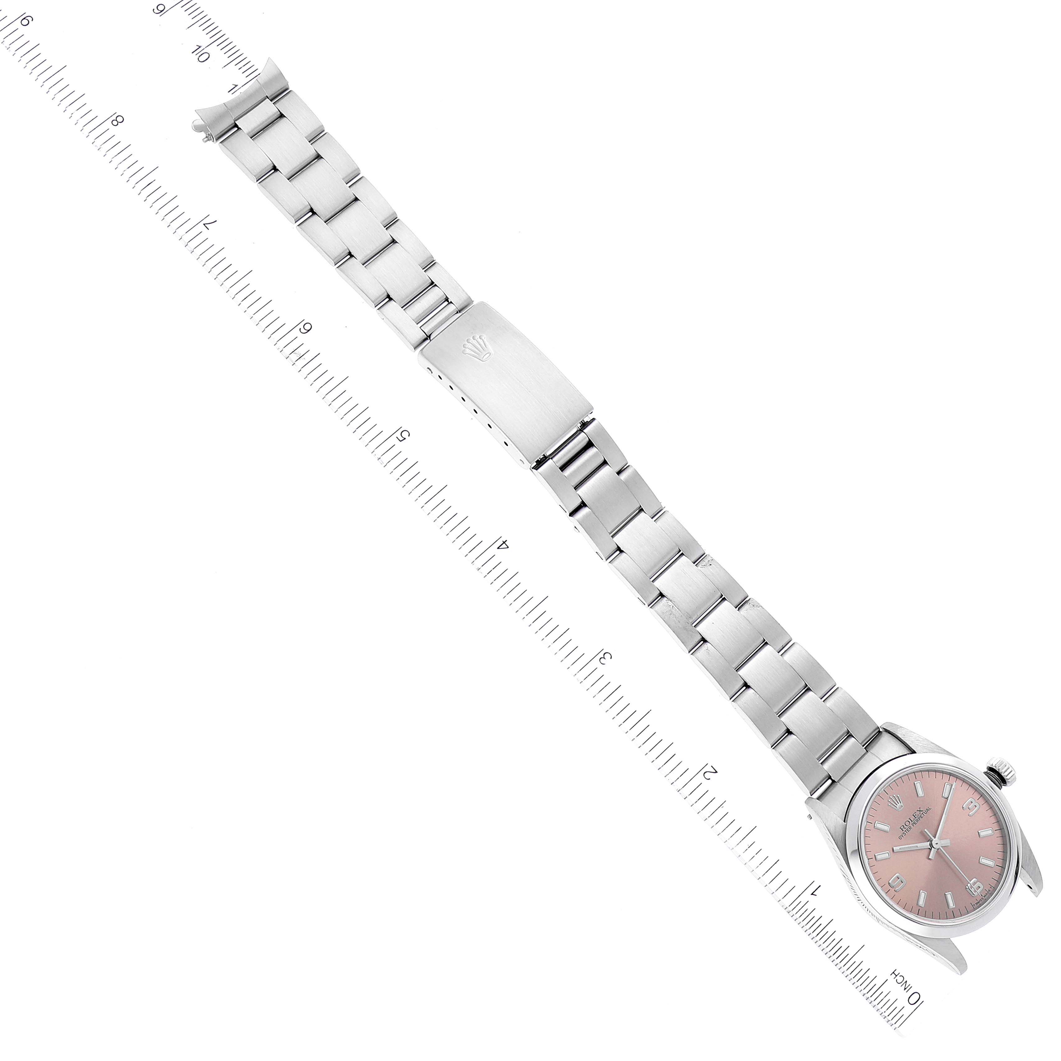The image shows a top-down view of a Rolex Mid-Size 77080 Women's Stainless Steel Pink Dial 77080 Women's Stainless Steel Pink Dial model watch with a pink dial and stainless steel bracelet clasp next to a ruler.
